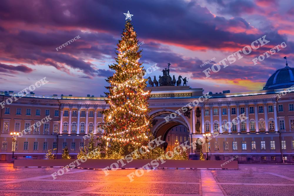 Фотография показывает Дворцовую площадь Санкт-Петербурга, украшенную к Новому году. Центральное место занимает ярко освещенная новогодняя елка, сияющая праздничными огнями. Арка Главного штаба гармонично вписывается в композицию, создавая эффект торжественности. На заднем плане видно красочное закатное небо, которое подчеркивает магию зимнего вечера в городе.